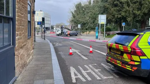 The rear of a police car on a road with two cones next to it. The is police tape down the road and track and car in the distance.