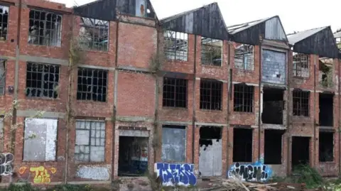 The derelict building has moss growing on it as has been daubed with graffiti. It has four storeys and bars on its windows which no longer have glass. 