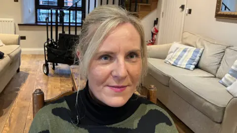 Sandra Larmour in a living room wearing a green and black jumper with her blonde hair tied up