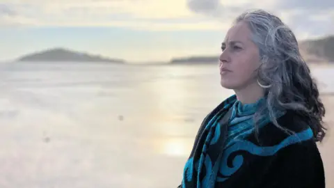 Parker A lady with grey and black hair looks out to the sea. She wears a striking blue and black patterned scarf. The shore is out of focus behind her as the sun shines. 