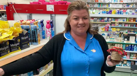 Justin Dealey Kerry Evans posing for a photo in Gadebridge Pharmacy in Hemel. She is wearing her uniform - a royal blue NHS coloured shirt with a collar with white edging and black cardigan. She is holding a jar of "Aunt Lucy's Marmalade" which has a Peruvian cloth on the top. She has highlighted blonde hair partly swept back into a clip and behind her are shelves of chemist goods.