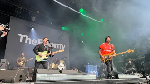 Band The Enemy are on stage performing. One is holding a green guitar singing into a microphone, while another in a red t-shirt is playing a bass. In the background is a screen reading: The Enemy. Monitor speakers are in the foreground on the front of the stage