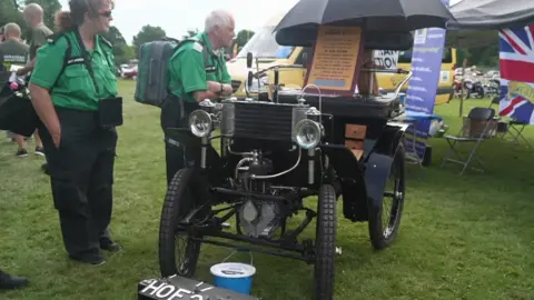 An early 20th-Century vintage car is on display with its exposed mechanical components, upright stance and narrow wheels, while a number plate reading “HOF2” sits beneath the front axle. 