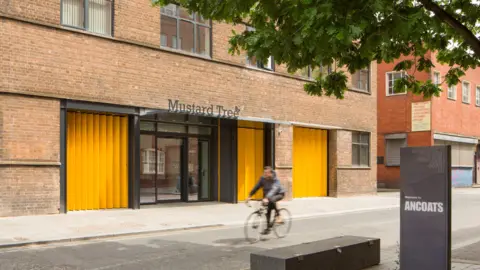 MustardTree A man cycles past a modern building advertising the charity 'Mustard Tree'.