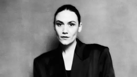 The Glasshouse A black and white photo of Nadine Shah. It is slightly out of focus and she is looking into the camera with pursed lips. She has dark hair which is scraped back  and is wearing a black blazer with large shoulders. She is standing in front of a grey background.