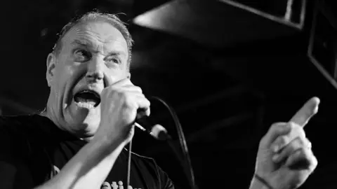 Getty Images Black and white image of Richard Jobson from The Skids whose mouth is wide open as he sings into a mic and points his finger 