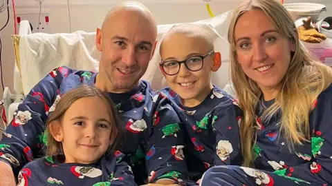 Fewster family A man with very short hair, a woman with long blond hair, a girl with medium-length brown hair and a boy with very short hair and glasses sit on a hospital bed. All are wearing blue Christmas pyjamas with festive pictures.