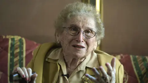 AFP via Getty Images Rosa Roisinblit smiles and gestures at the camera during an interview with AFP in Buenos Aires, 2016. She wears a pale mustard jacket and matching shirt, holding a walking cane in her right hand. Her nails are painted pink, and she wears wire-rimmed spectacles. 