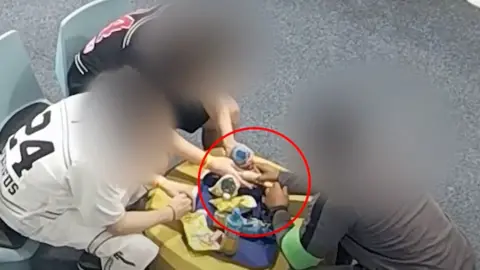 A still from a CCTV camera showing three people sat on blue chairs around a low yellow table. They are leaning into the table, which has a blue canteen tray on it. One person can be seen handing a package to the person sitting opposite him.