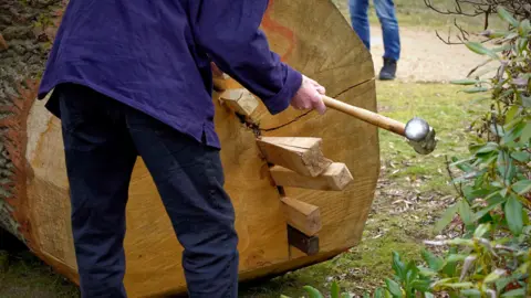 The Sutton Hoo Ship's Company Man hammering wooden pegs into the base of an oak tree trunk to split it