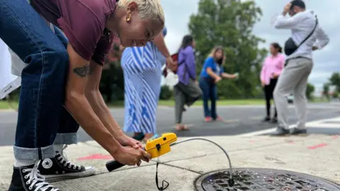 HELEN GEBREGIORGIS A woman holding a yellow test device measures gases emitted by the street drainage