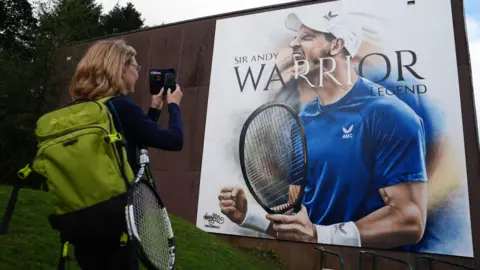 Woman carrying a green backpack and a tennis racket uses her phone to take a photograph of a large artwork of Andy Murray. It is a picture of Murray in a blue t-shire and white cap. It says Andy Murray. Warrior. Legend. 