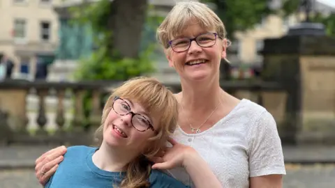 A young girl smiles with her head cocked to the side as she leans back against her mother, who is smiling and has her arm wrapped around her. The pair are stood outside.