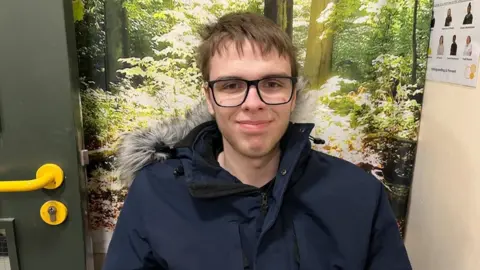 Ollie Conopo/BBC Young man with short dark hair and glasses wearing a hooded coat