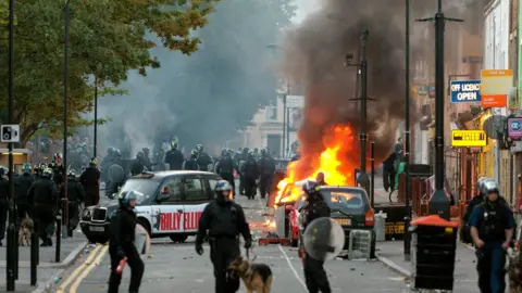 Getty Images Riot police look on as a car burns