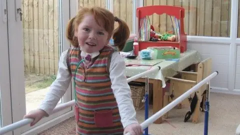 John Huggins Sophie, with red hair in bunches and wearing a striped dress with a white long-sleeved T-shirt, stands holding white physiotherapy training bars. She has a tube coming from her throat and is smiling at the camera. In the background is a play table with a puppet theatre, next to a child's chair. She is in a conservatory.