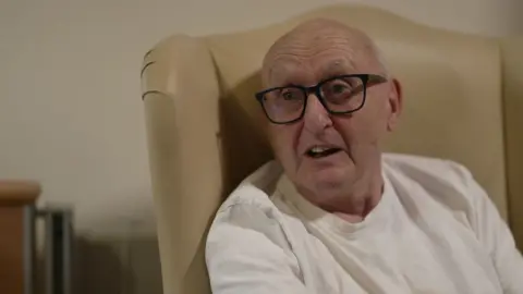 Older man sitting in an armchair wearing glasses and a white T-shirt. The chair is cream in colour and his glasses are black-framed.