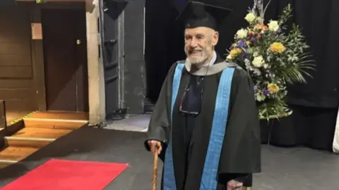 Kingston University A man walking across a graduation stage. He is wearing a gown with a blue tab and a black cap. He holds a walking stick. There is a large bunch of flowers in the background.