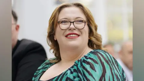 A woman in a black and green striped dress smiles while looking into the distance. She is wearing glasses.