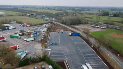 National Highways Drone shot of a new tarmacked lorry parking area, with fields and some industrial buildings nearby.