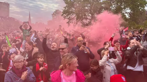 York City fans cheer on their team as an open top bus passes. Red flare smoke fills the air.
