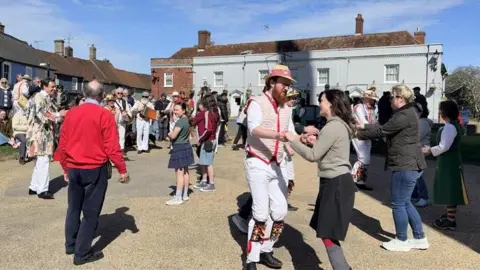 Jamie Niblock/BBC A crowd of people getting involved in Morris dancing in a town square.
At the front of the picture is a Morris man holding hands with a member of the public as they dance. There are old buildings in the background.