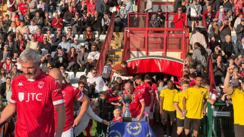 Players come out of the tunner and go past the trophy as fans in the background cheer on from the stand.