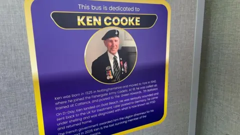 Olivia Richwald/BBC A purple plaque on a grey wall reads 'This bus is dedicated to Ken Cooke'. It has details about Ken's life written underneath and an image of an old man wearing a military uniform and medals. 