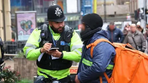 A police officer in high vis clothing takes notes while he talks to a food delivery rider who's face is blurred in the image.