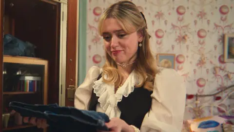 Netflix Aimee Lou Wood as Aimee in Sex Education. Aimee, a young white woman, has long blonde hair styled half-up-half-down with a short fringe to the side of her face. She wears a black waist coat over a frilly cream shirt and a pearl choker-style necklace. Aimee is pictured inside a bedroom with decorative pink and white wallpaper and holds a pair of jeans in front of her.