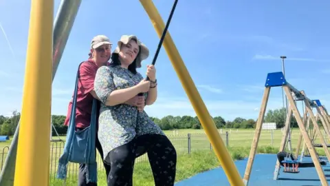A young woman is on a swing. She is wearing a sun hat and looks happy. A woman is standing behind her, smiling. It's a sunny day and both women look happy.