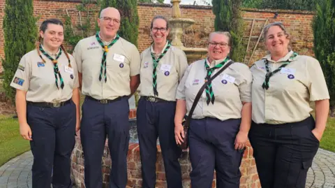 Five cub scout leaders - three women and one man - standing next to each other wearing their uniform of beige tops and navy trousers. They are all smiling at the camera. They are outside in a garden and there is a water fountain behind them.