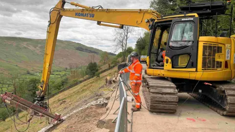 A large digger installs a metal rod in the slope near Kirkstone Pass. The yellow digger is on the road and a person wearing orange fluorescent uniform watches over. 