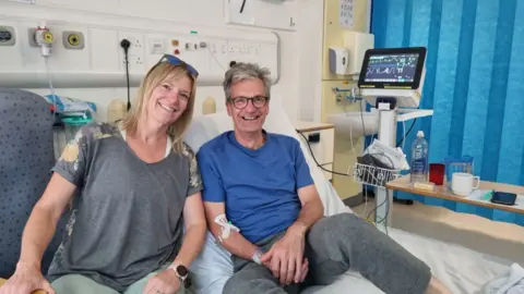 BBC A woman sitting next to a man on a hospital bed