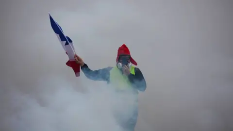 Getty Images A demonstrator holds a French flag as he walks amid the tear gas during a protest of Yellow vests (Gilets jaunes) against rising oil prices and living costs, on December 1, 2018 in Paris
