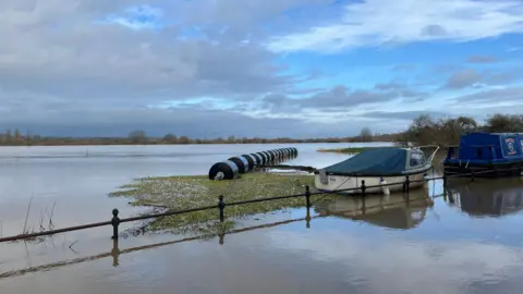 A large, countryside landscape is flooded. Metal posts are submerged in water. Boats are floating, as well as bales of hay in black bags.