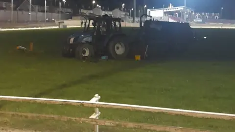 A blue tractor sits in the middle of a dog racing track in Derry. White fencing can be seen in the foreground, and a spectator's stand is visible in the background. It is night time and the stadium is flood lit.