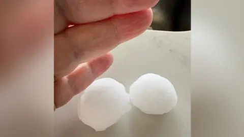 A person's middle, ring and baby finger next to two large golf ball-sized white hail stones on a white surface.