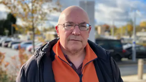 Shariqua Ahmed/BBC Mark Keen is stood in the middle of the image looking at the camera. He is wearing glasses, an orange jumper and black coat. 