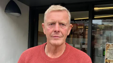 Hugh Smy has short grey hair and is wearing an orange jumper. He is standing in a shop front.