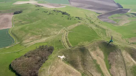 Getty Images An aerial image of a hillside which has a large stone obelisk in one corner and a white horse etched onto the hillside in another corner