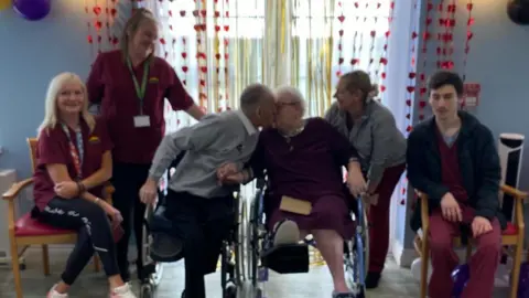 Susan Salvatore-Davies Staff at Picton Care Home in Porthcawl are wearing burgundy tops. They are sat and standing around Colin, who is wearing a grey jumper and white shirt, and Pamela. She is wearing a purple dress. 