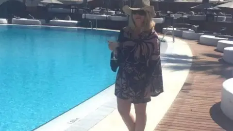 Gemma Louis Gemma beside a very blue pool holding a glass of wine. It is very sunny and she is wearing a purple mini dress, sunglasses and a beige cowboy hat. 