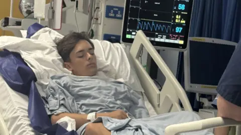 A teenager in a hospital bed attached to a monitor with his eyes closed