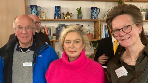 Daniel Mumby A man in a blue coat is stood next to two women. One woman, Tiverton and Minehead MP Rachel Gilmour, has a pink jumper, the other woman is wearing a brown shirt.