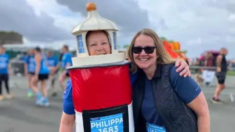 A woman dressed as a white and red lighthouse. She is side hugging a woman wearing a blue vest and blue shirt. Behind them are other runners. 