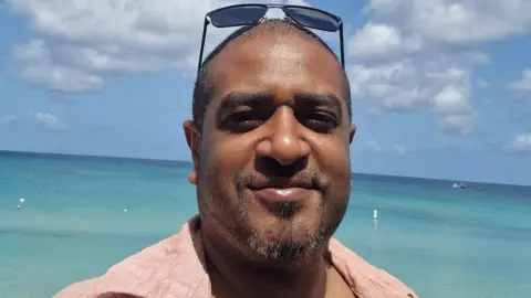 A man wearing sunglasses on his head and smiling at the camera. He is wearing a shirt and standing on the beach. He looks happy 
