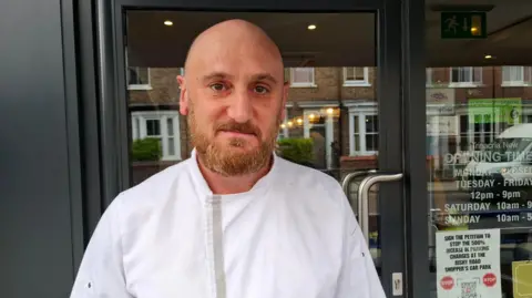 Beppe Lombardo has a bald head and a ginger beard. He is wearing a chef's uniform and is standing outside a restaurant door.