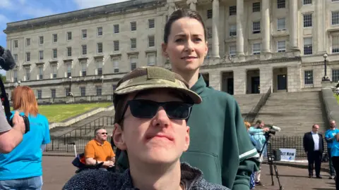 Tiernan Flood wears a cap and sunglasses while he and his mother, Tanya Davis, pose for a photo in front of Stormont