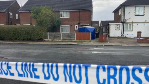 The front of a semi-detached house with a blue forensic police tent in the front yard. Blue and white police tape is visible in the foreground